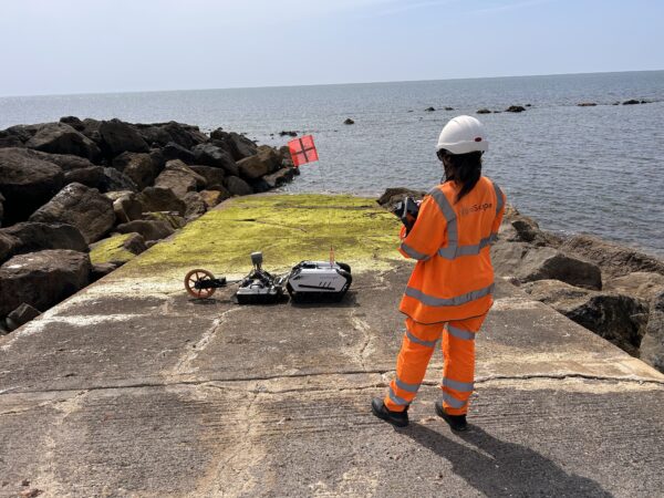 Remote-controlled tracked machine towing GPR equipment across slippery coastal defences.